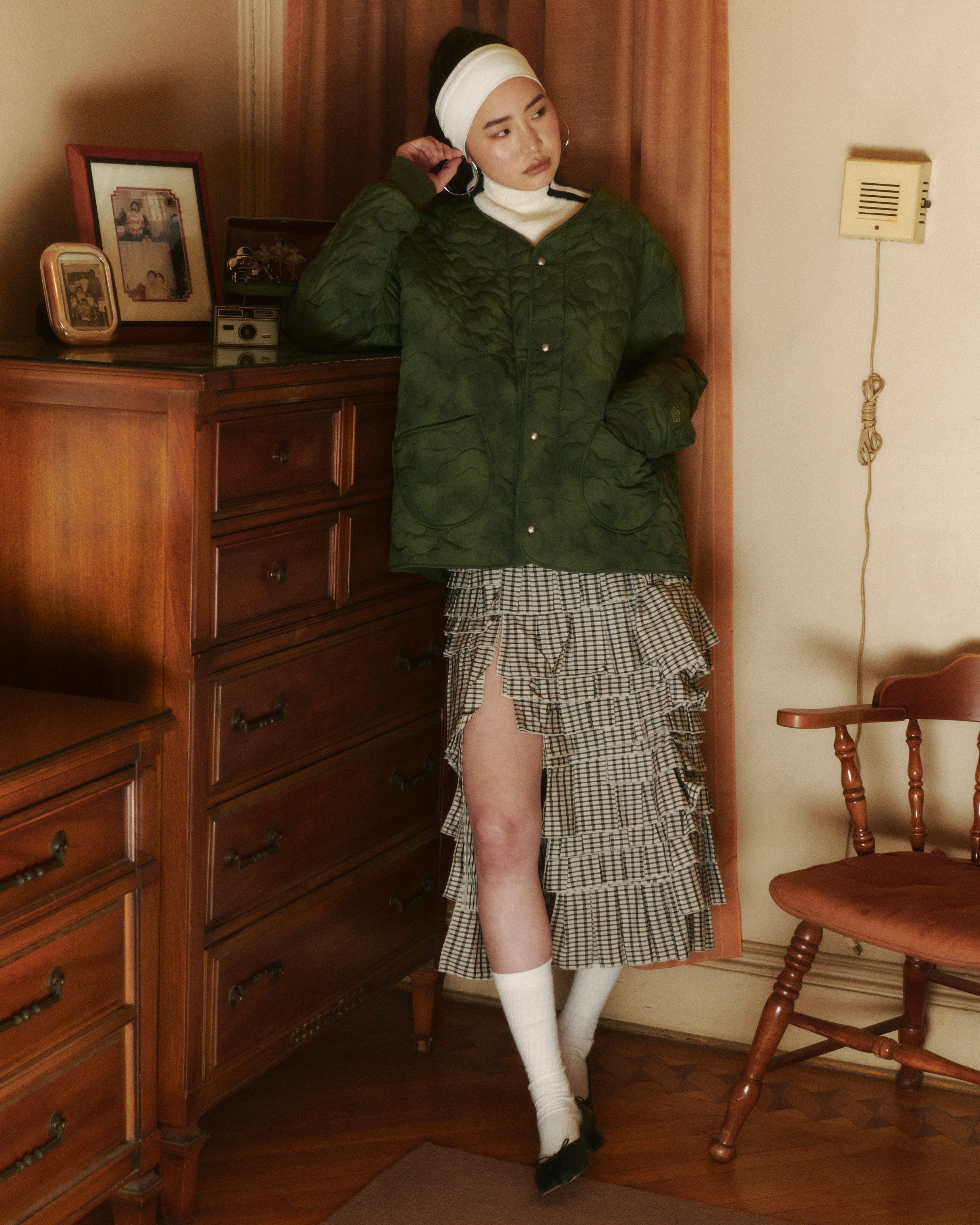 A woman wearing Sundae School's MATCHA MOCHI QUILTED MILITARY LINER, a houndstooth ruffled skirt with a high slit, white socks, black shoes, and a white headband leans against a wooden dresser in a retro-style vintage room.