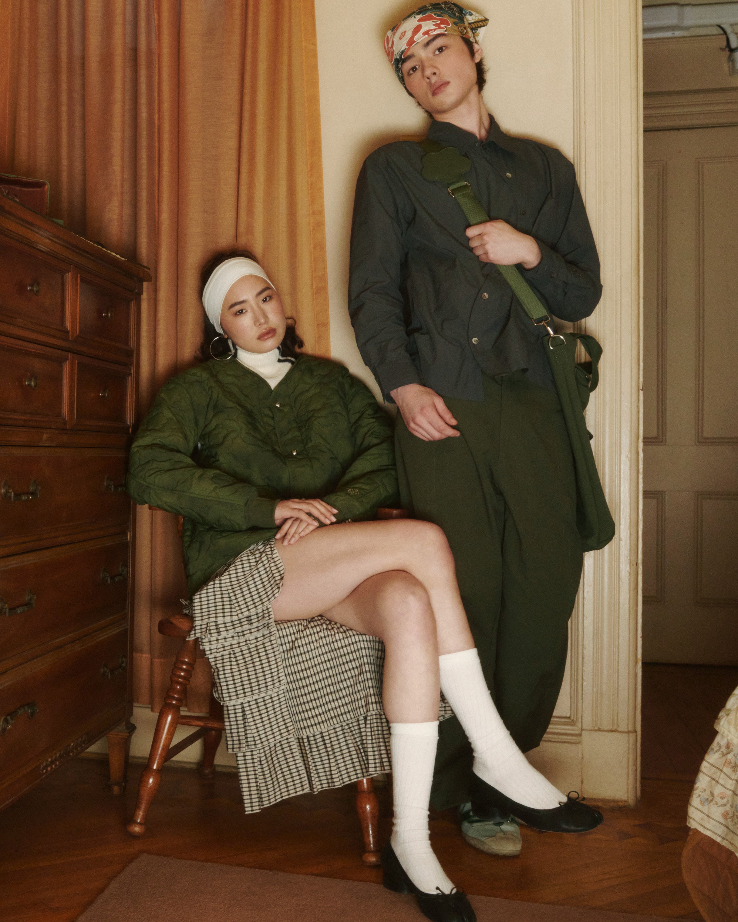 Two people pose indoors; one sits on a wooden chair in Sundae School’s MATCHA MOCHI QUILTED MILITARY LINER, paired with a checkered skirt, white headband, and black shoes, while the other stands nearby in a transitional jacket, dark pants, and colorful accessories.