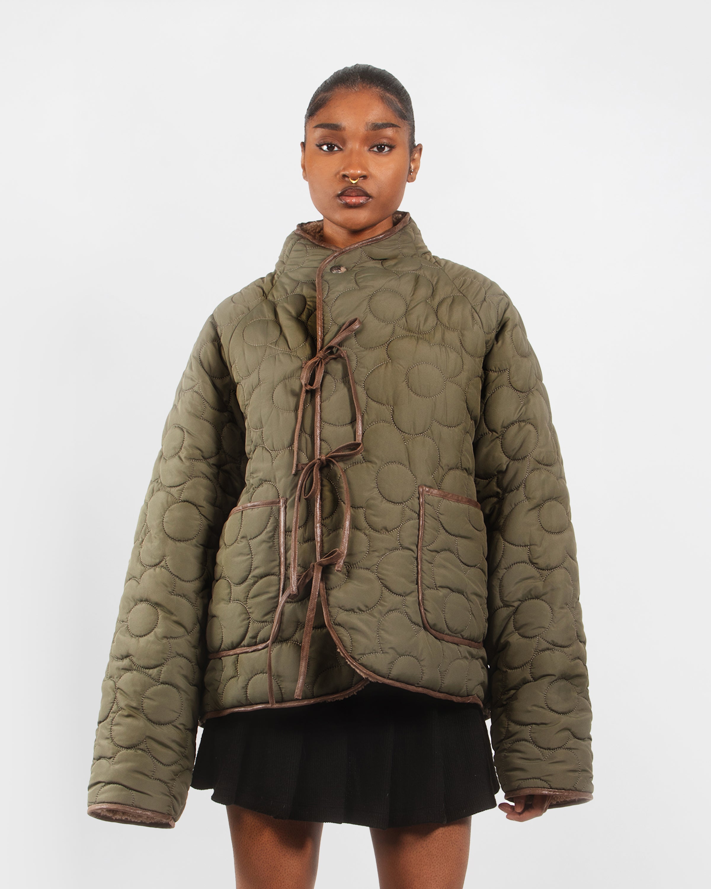 A woman wears the Sundae School REVERSIBLE FLOWER QUILTED JEOGORI FLEECE—an oversized, olive-green puffer jacket with large pockets and brown trim—paired with a black skirt against a plain white background.