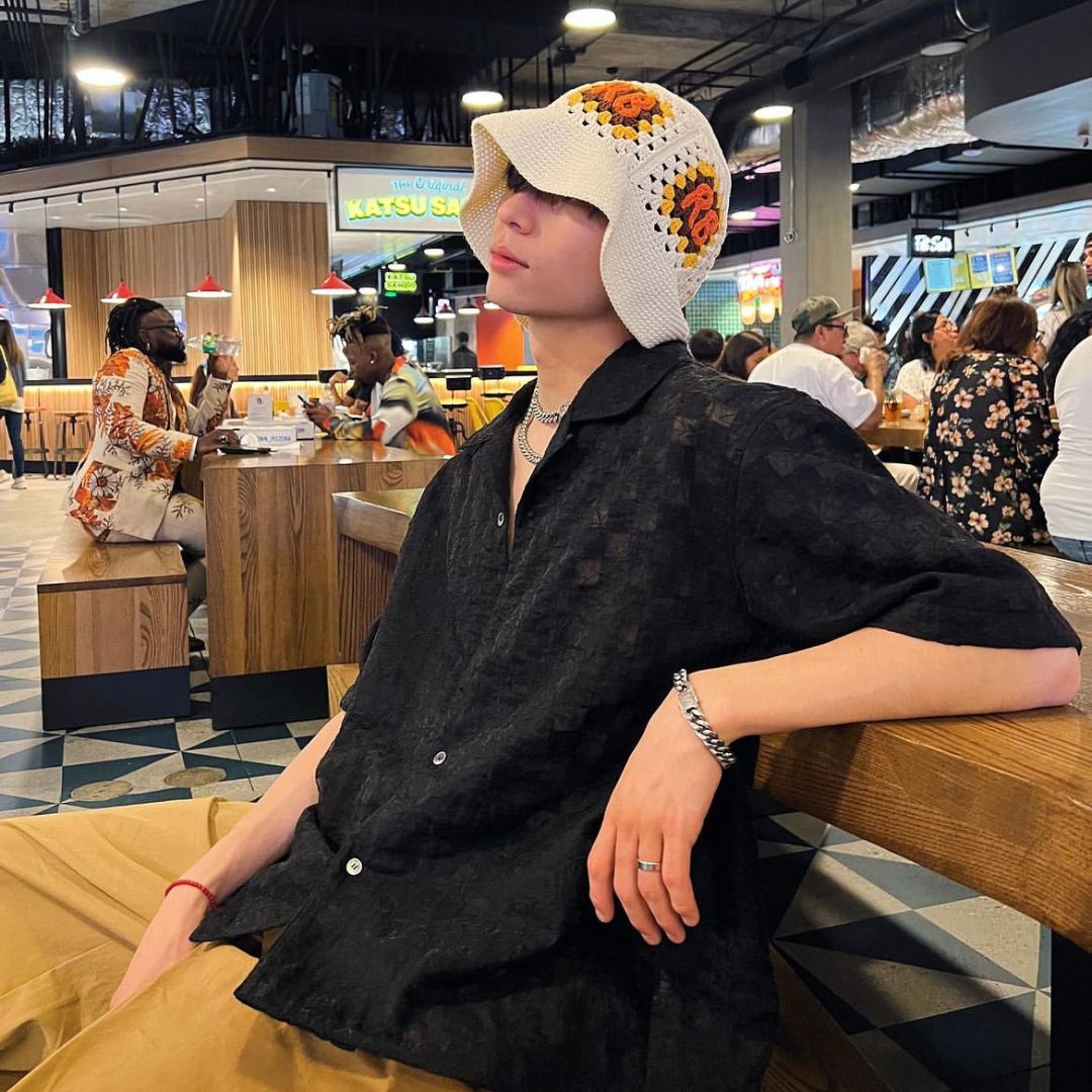 A person lounges indoors at a food court wearing a textured black button-up shirt and tan wide-leg pants. They accessorize with a chunky chain necklace, silver jewelry, and a crocheted bucket hat with orange flower designs. The ambiance is trendy and social.