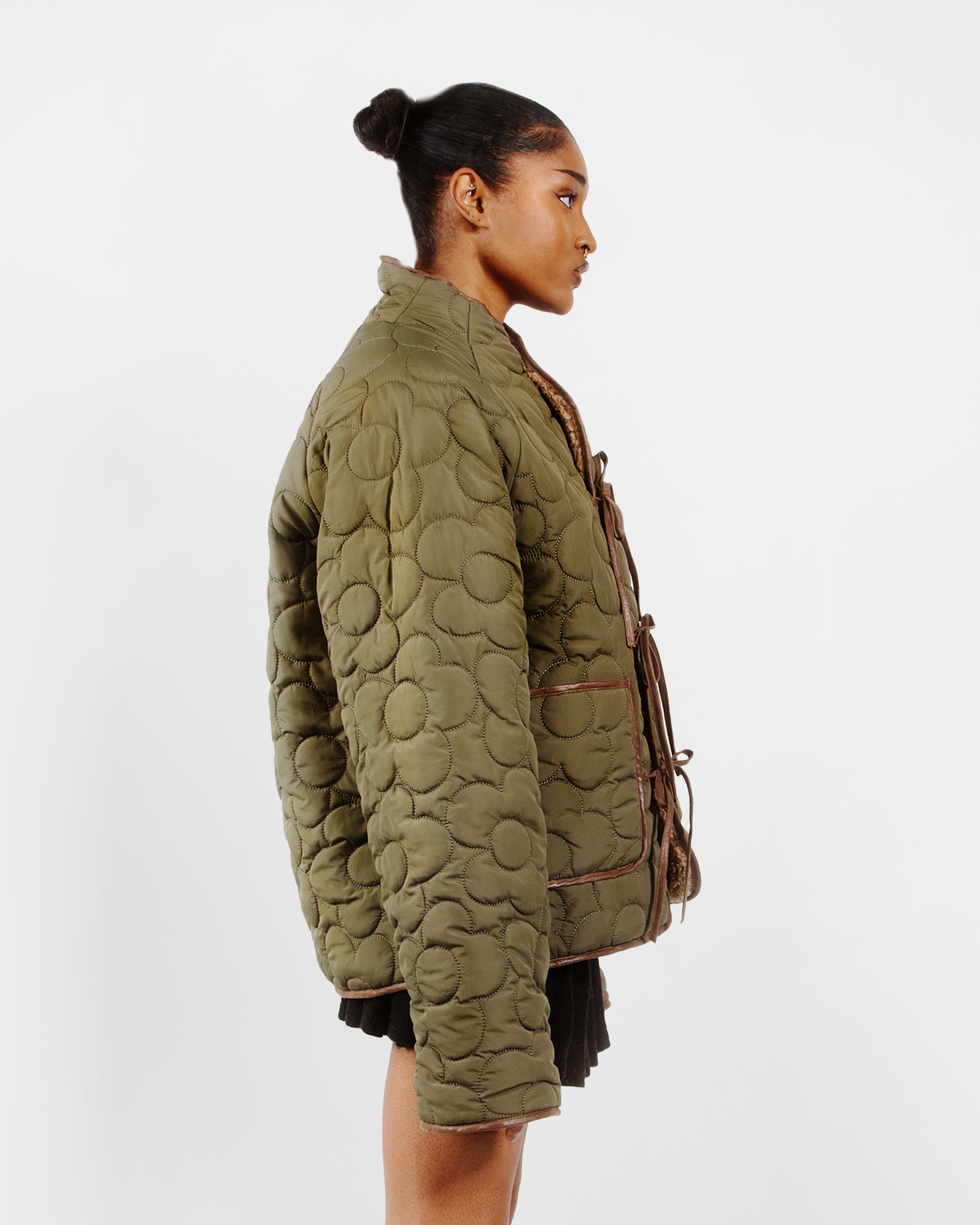 A person with their hair in a bun is shown in profile wearing Sundae School’s Reversible Flower Quilted Jeogori Fleece, an oversized olive-green jacket with large circular patterns and front ties, paired with a black skirt against a plain white background.