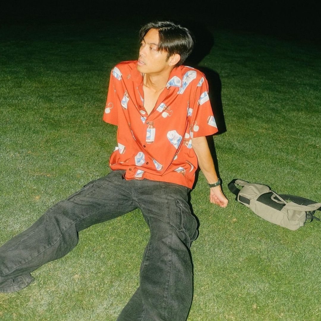 This individual sits on the grass at night wearing a bright red short-sleeve button-up shirt printed with bottles and cherries, paired with black baggy pants. They are lit by a strong flash, giving the photo a candid, vintage feel. A small olive green bag rests beside them.