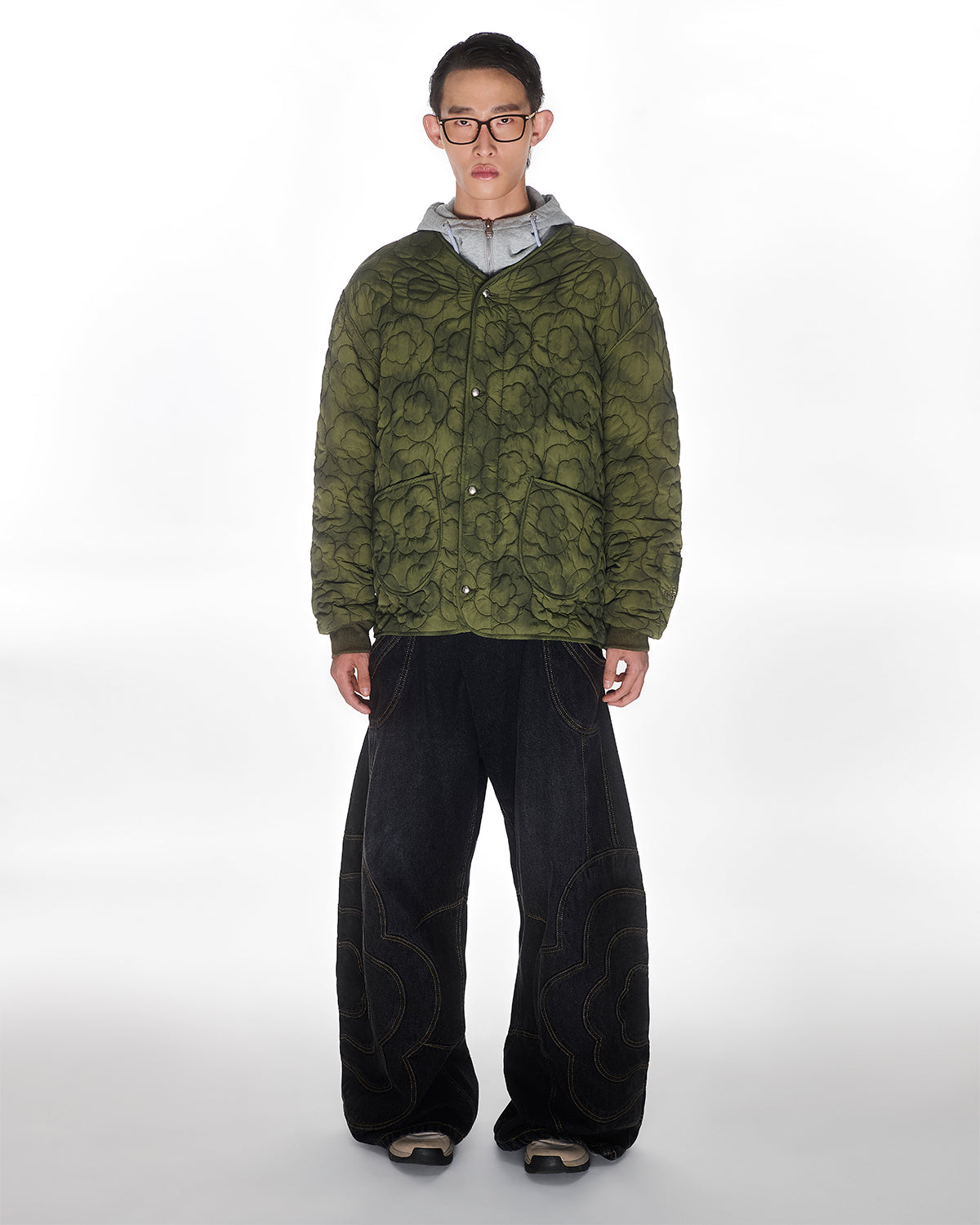 A person stands facing forward in glasses, a green quilted jacket with a mochi flower appliqué over a grey hoodie, Sundae School BLACK MOCHI PANELED PANTS (x JOONIVERSE), and sneakers, against a plain white background.