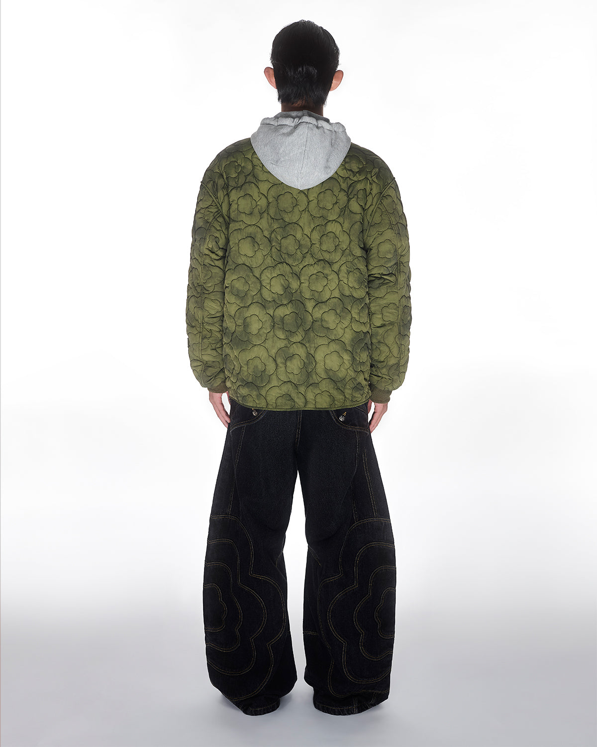 A person stands with their back to the camera, wearing a green floral-patterned quilted jacket over a gray hoodie, paired with Sundae School BLACK MOCHI PANELED PANTS (x JOONIVERSE). The background is plain white.
