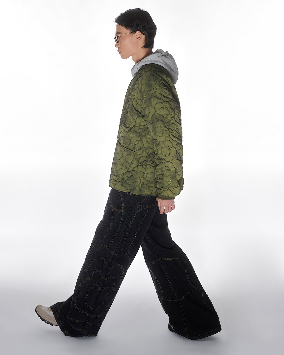 A person in sunglasses and beige shoes walks sideways against a white background, wearing the Sundae School MATCHA MOCHI QUILTED MILITARY LINER—a green floral transitional jacket—over a gray hoodie with wide-leg dark jeans featuring large stitched designs.