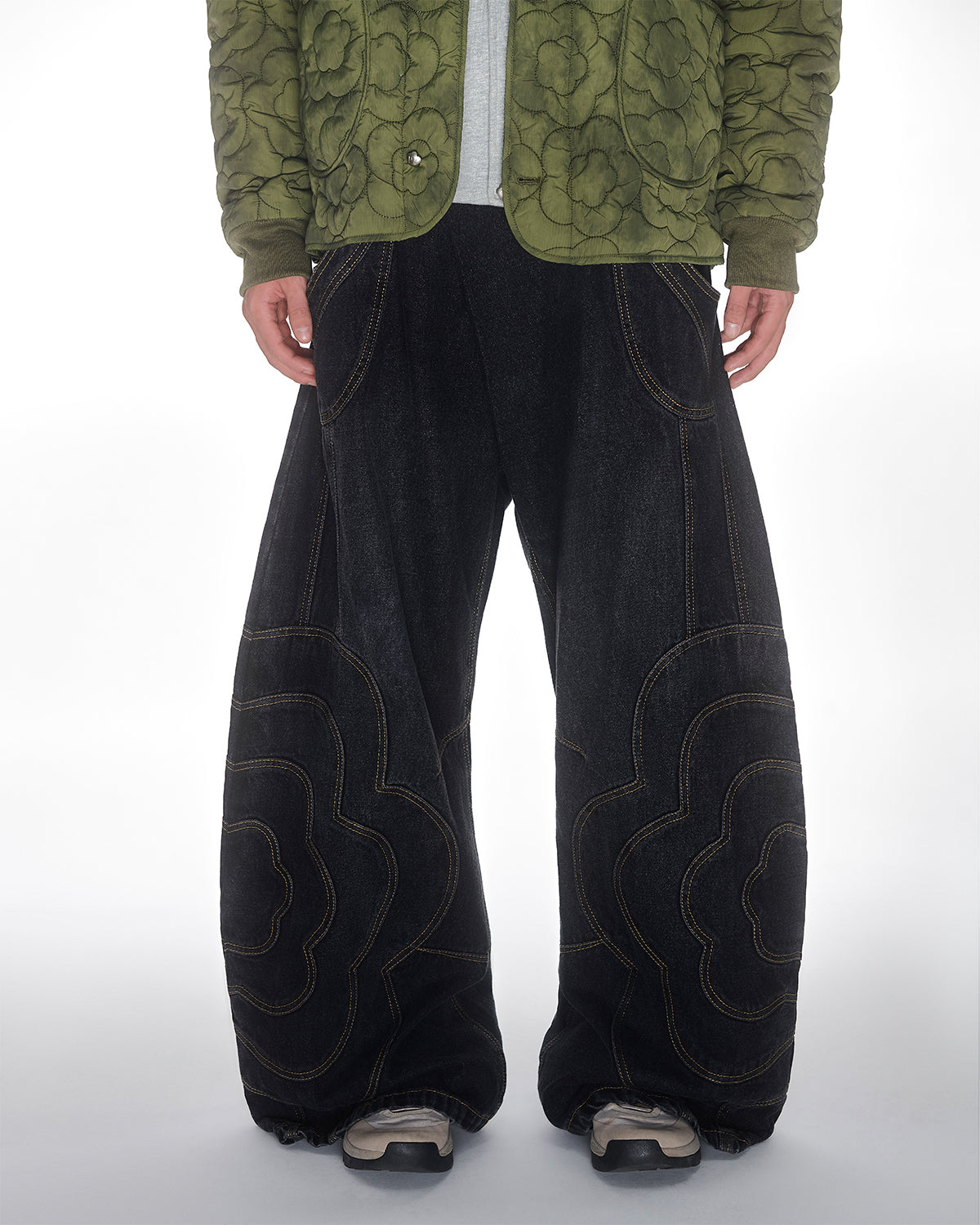 A person wears Sundae School's BLACK MOCHI PANELED PANTS (x JOONIVERSE), unisex wide-leg denim jeans with large wavy yellow mochi flower appliqué, paired with a green quilted jacket and beige sneakers against a white background.