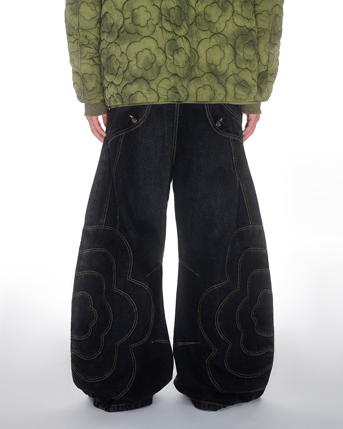 A person faces away, showcasing Sundae School’s BLACK MOCHI PANELED PANTS (x JOONIVERSE) paired with a green patterned jacket, highlighting the unisex design against a plain white background.