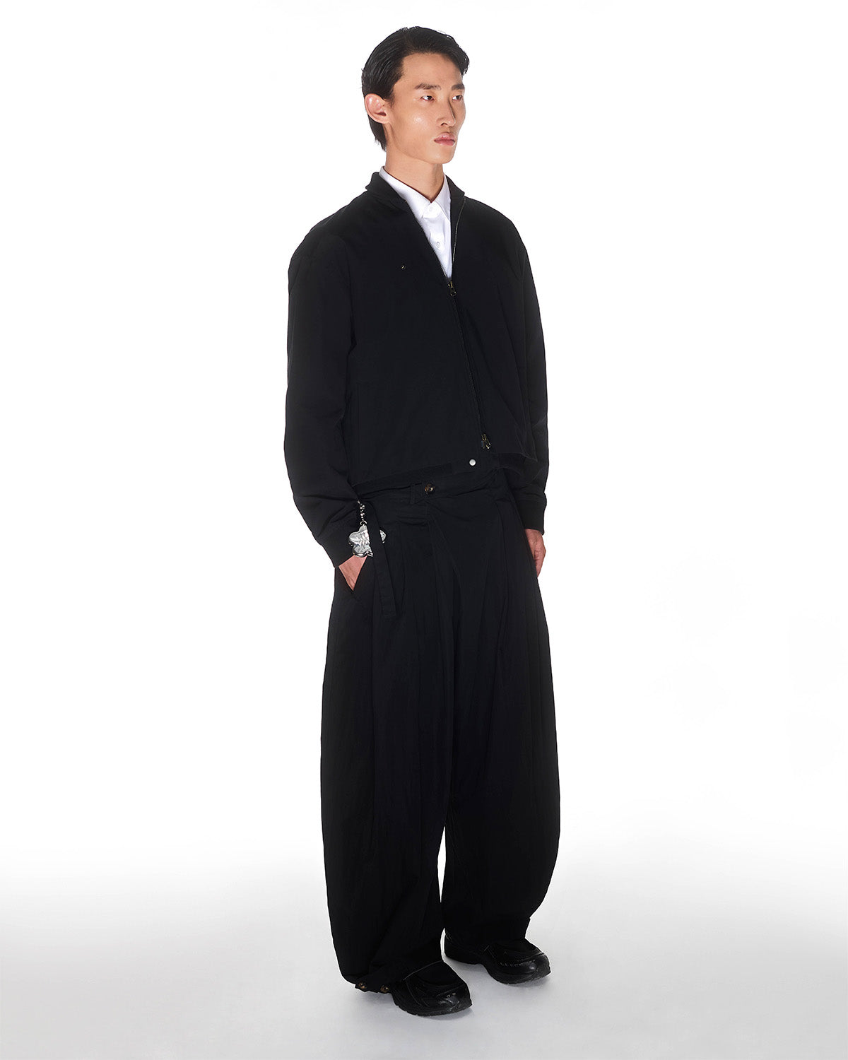 A person with short dark hair stands against a white background wearing Sundae School BLACK PLEATED NYLON PANTS, a black bomber jacket over a white shirt, and black shoes, with their hands in their pockets.