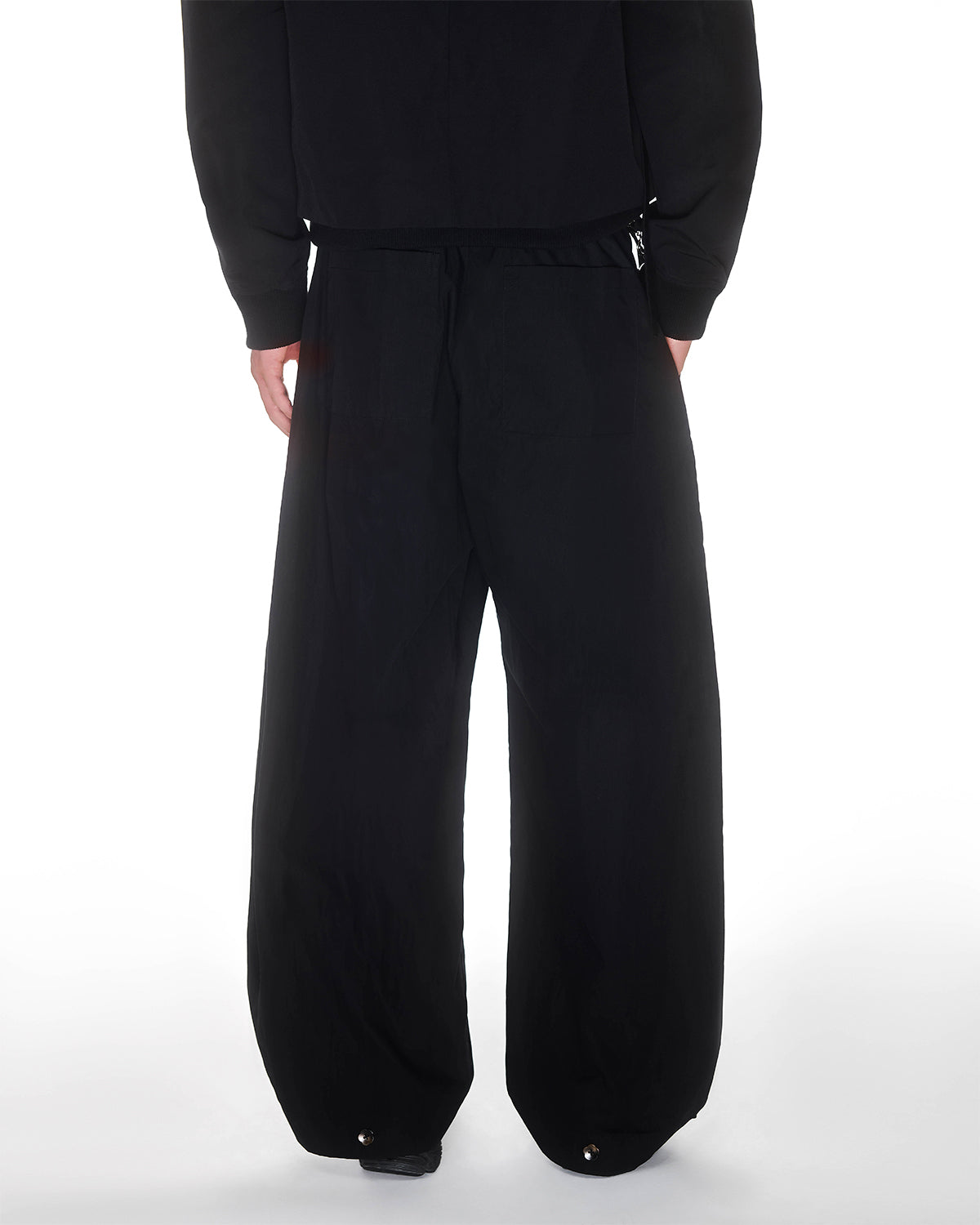 A person wearing Sundae School BLACK PLEATED NYLON PANTS and a black top stands with their back to the camera against a white background.