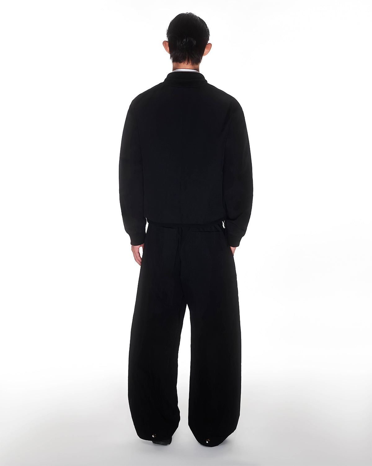 A person with dark hair faces away, wearing a loose black jacket and Sundae School’s BLACK PLEATED NYLON PANTS against a plain white background.