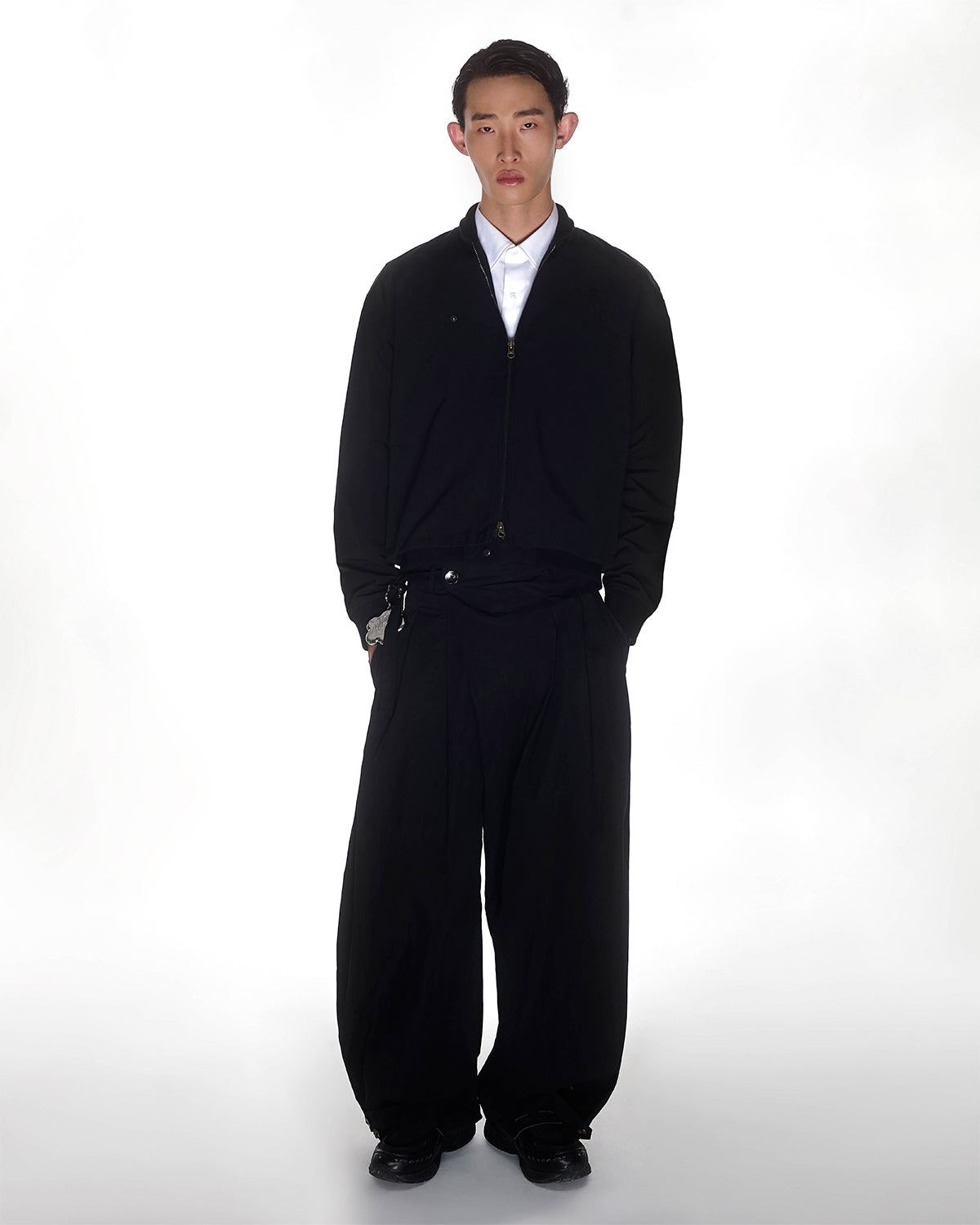 A young man stands facing forward against a white background, wearing Sundae School BLACK PLEATED NYLON PANTS with an adjustable D-ring belt, a black zip-up jacket, white collared shirt, and black shoes. His hands are in his pockets and he has a neutral expression.