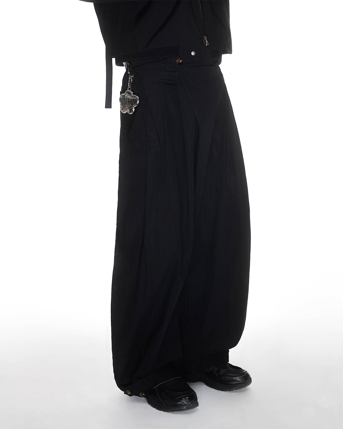 A person models Sundae School BLACK PLEATED NYLON PANTS against a white background, paired with black shoes and a black jacket featuring a keychain on the waistband. Only the lower body is visible.