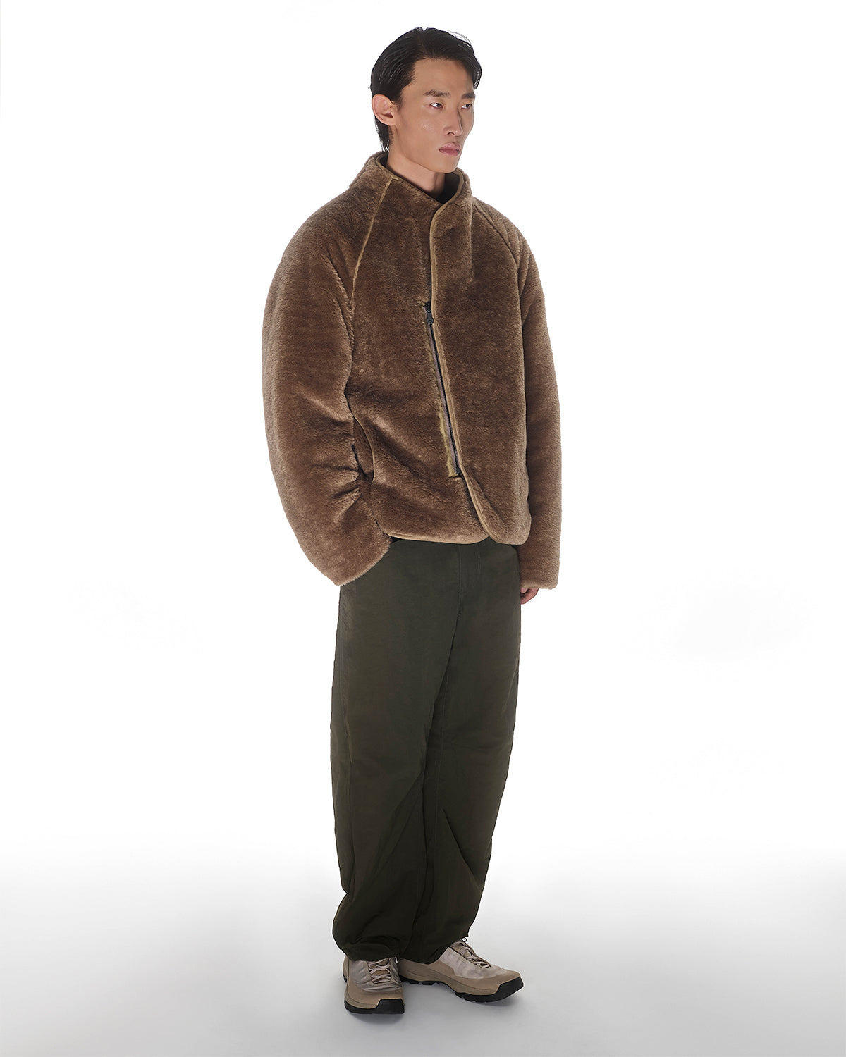 A man with short dark hair and a neutral expression stands against a white background, wearing Sundae School's REVERSIBLE FLOWER QUILTED JEOGORI FLEECE, olive green pants, and beige sneakers, with his hands relaxed at his sides.