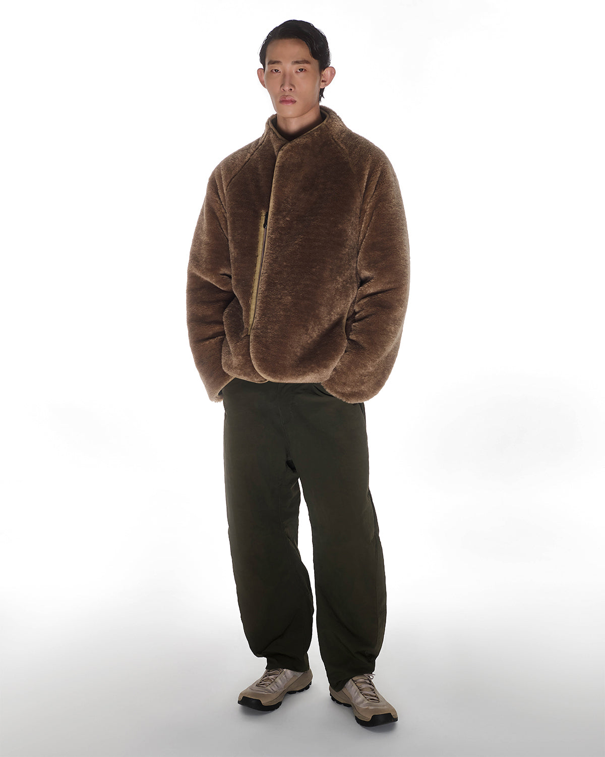 A person stands against a white background wearing the Sundae School REVERSIBLE FLOWER QUILTED JEOGORI FLEECE, paired with dark green pants and beige sneakers. They have short dark hair, a neutral expression, and hands in the jacket pockets.