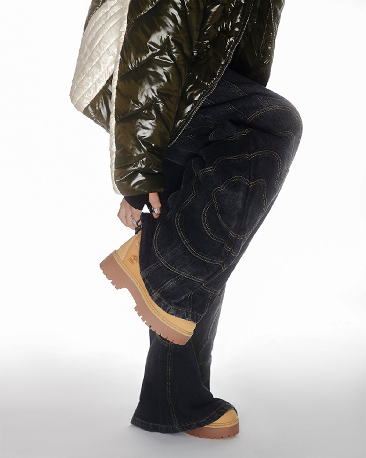 A person wears Sundae School BLACK MOCHI PANELED PANTS (x JOONIVERSE), tan platform boots, and a green quilted jacket, shown waist down, standing on one leg while adjusting the other shoe.