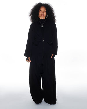 A person with curly hair wears wide-leg black pants, layered necklaces, hoop earrings, and the Sundae School QUILTED BOLO TIE CARDIGAN with a 떨 logo bolo tie, standing against a plain white background.