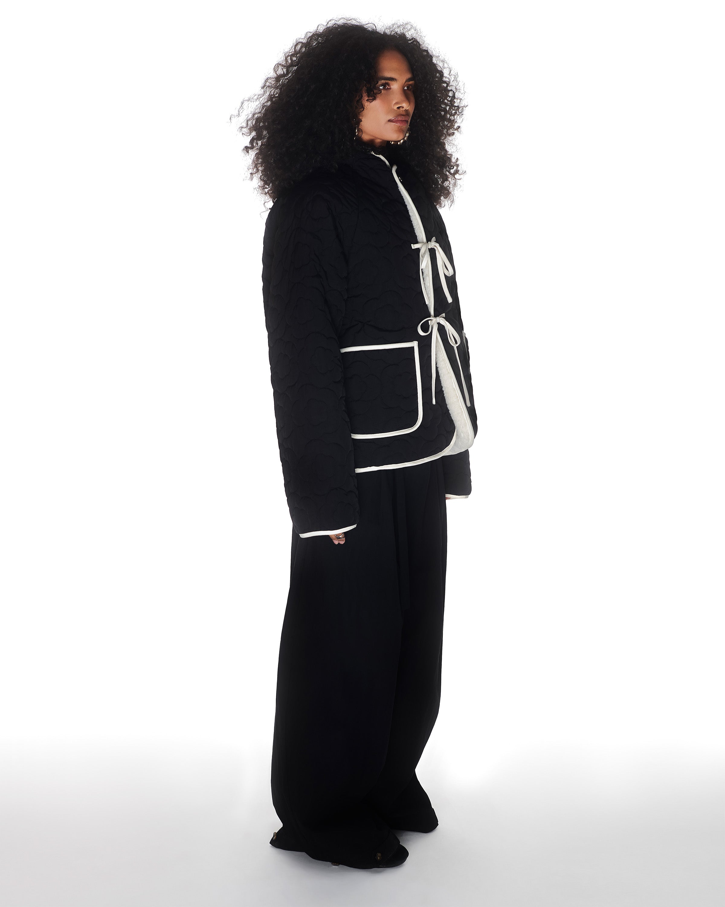 A person with long curly hair wears the Sundae School BLACK IVORY REVERSIBLE FLOWER QUILTED JEOGORI FLEECE, styled with loose black pants, standing against a white background.