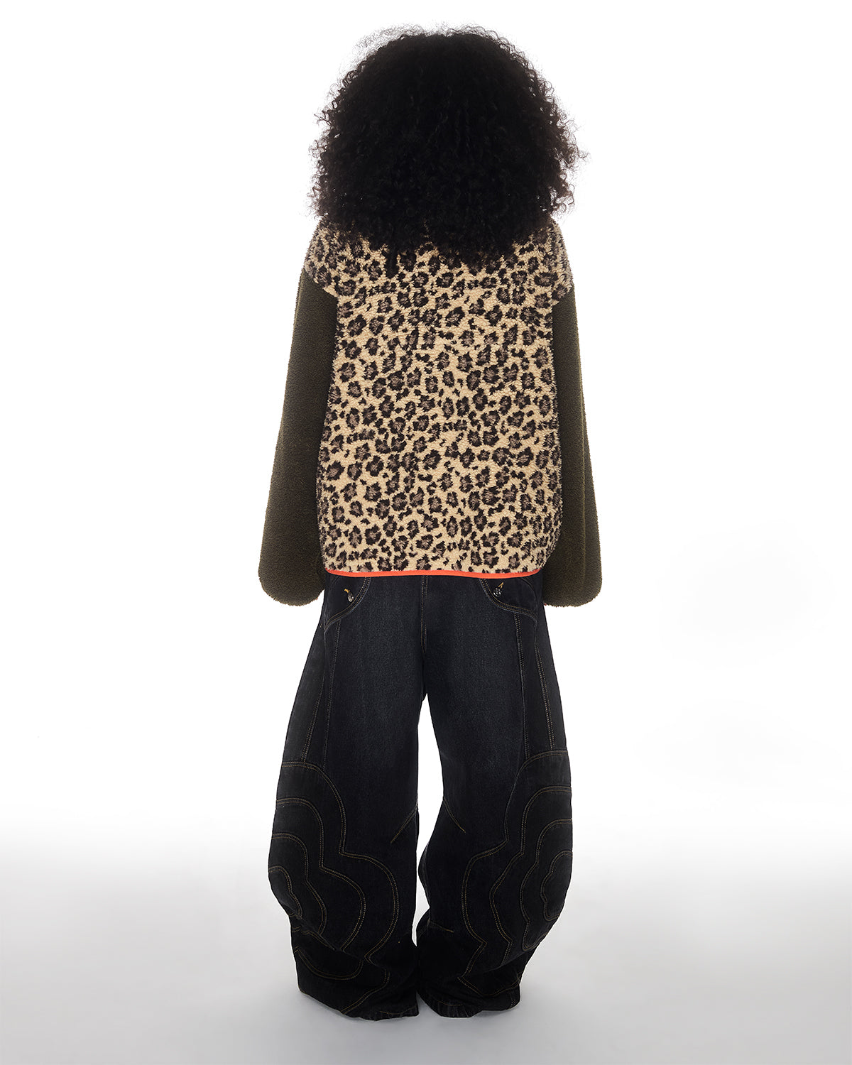 Facing away, a person with curly hair wears the Sundae School Tiger Mom Fleece—a sherpa jacket with fuzzy leopard print and olive green sleeves—paired with loose dark denim jeans, all set against a plain white background.