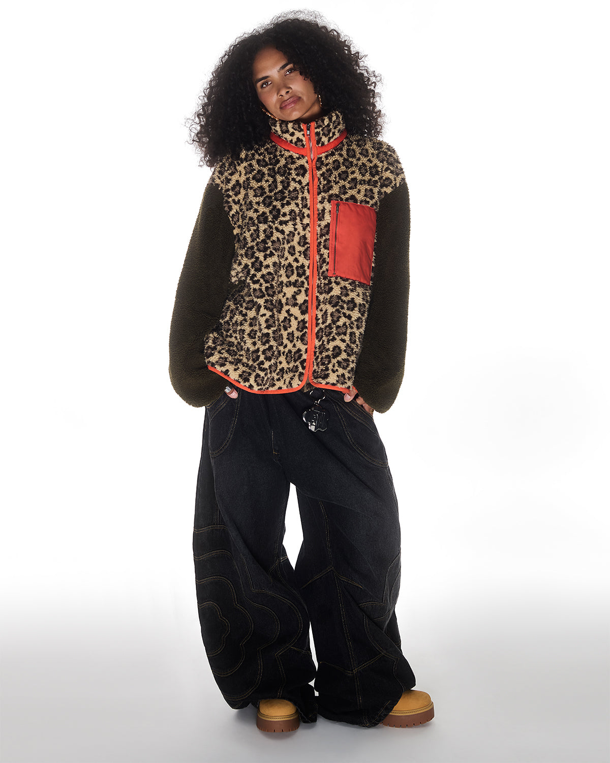 A person with curly hair wears a leopard print jacket with a red pocket, Sundae School BLACK MOCHI PANELED PANTS (x JOONIVERSE), and tan shoes, standing against a white background.