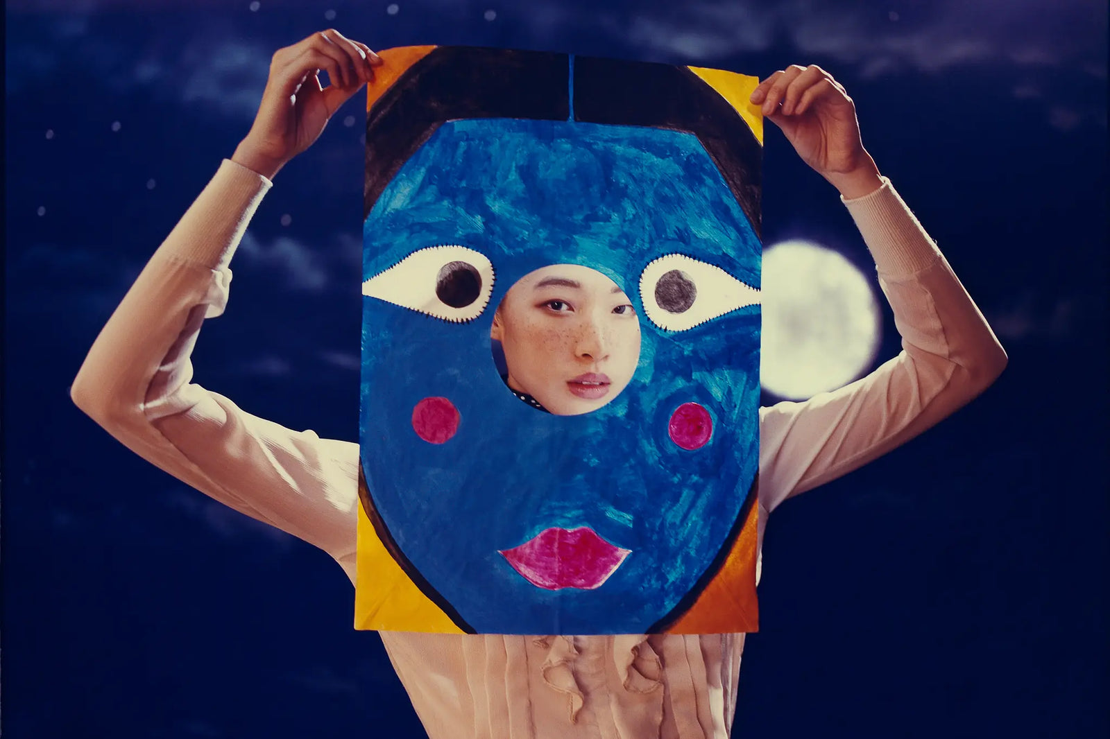 A person holding up a large, colorful art print or poster featuring a stylized blue face with pink cheeks, white eyes, and a cutout of their own face in the center, set against a dark night sky with a full moon.