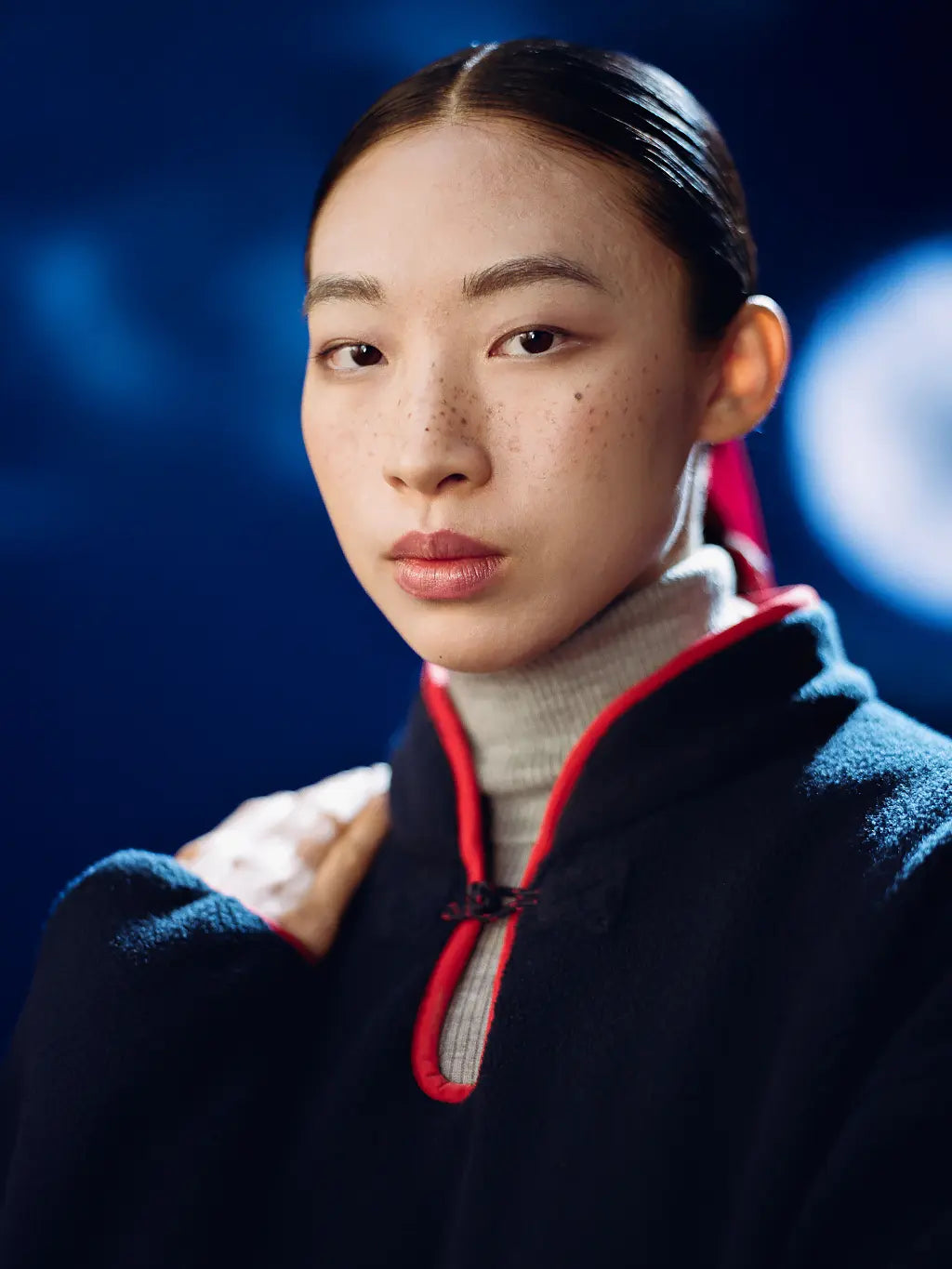 A close-up portrait of a model wearing Sundae School Navy Teardrop Knot Fleece with red trim over a light gray turtleneck, set against a dark blue background with soft bokeh lights.