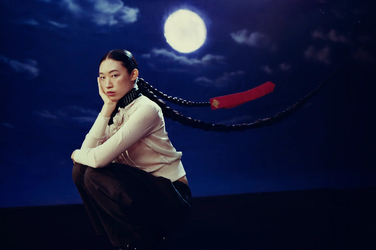 A person crouches in a dramatic pose against a night-sky backdrop with a full moon and clouds. The person wears a light beige top and dark pants, with a long black braided hair extension or wig featuring a bright red end piece.