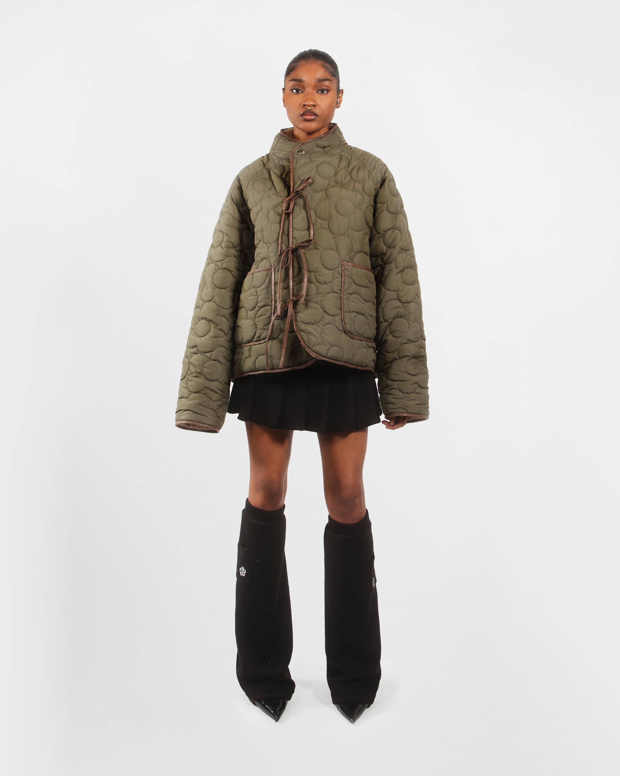 A person stands against a plain white background wearing Sundae School's REVERSIBLE FLOWER QUILTED JEOGORI FLEECE, an oversized green teddy sherpa coat with large front pockets, paired with a black skirt and tall black leg warmers over pointed shoes.