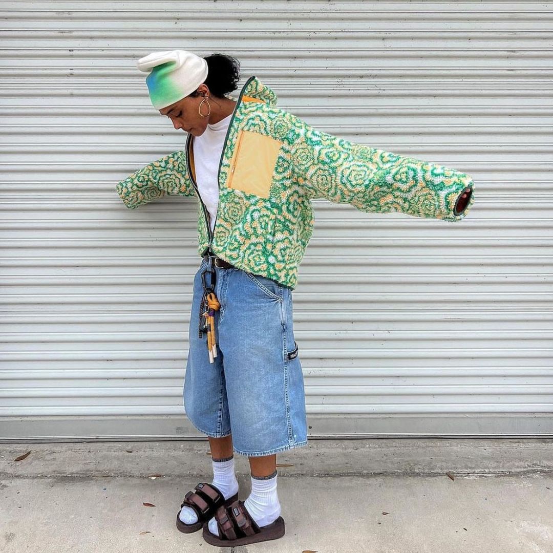 Standing against a corrugated metal wall, this person wears a fuzzy, green-and-yellow floral zip-up jacket with an oversized yellow chest pocket. They have on long denim shorts, sandals with socks, large hoop earrings, and a pastel beanie. Keys hang from their belt loop, adding flair.