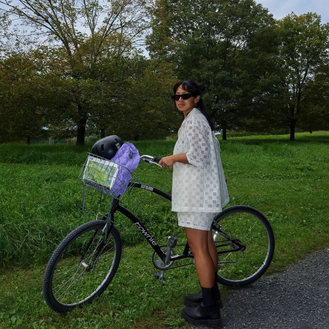 A person pauses beside a black cruiser bicycle in a grassy, tree-lined park. They wear a coordinated cream-colored mesh-textured set—boxy top and shorts—paired with chunky black boots and dark sunglasses. A lavender puffer bag sits in the bike’s basket.