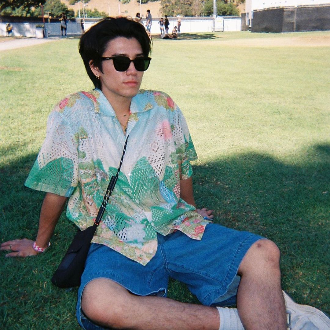 A person sits on the grass in a sunny park, wearing a loose pastel floral mesh button-up shirt and baggy denim shorts. They have dark hair, black sunglasses, a black shoulder bag, and a casual, relaxed posture.