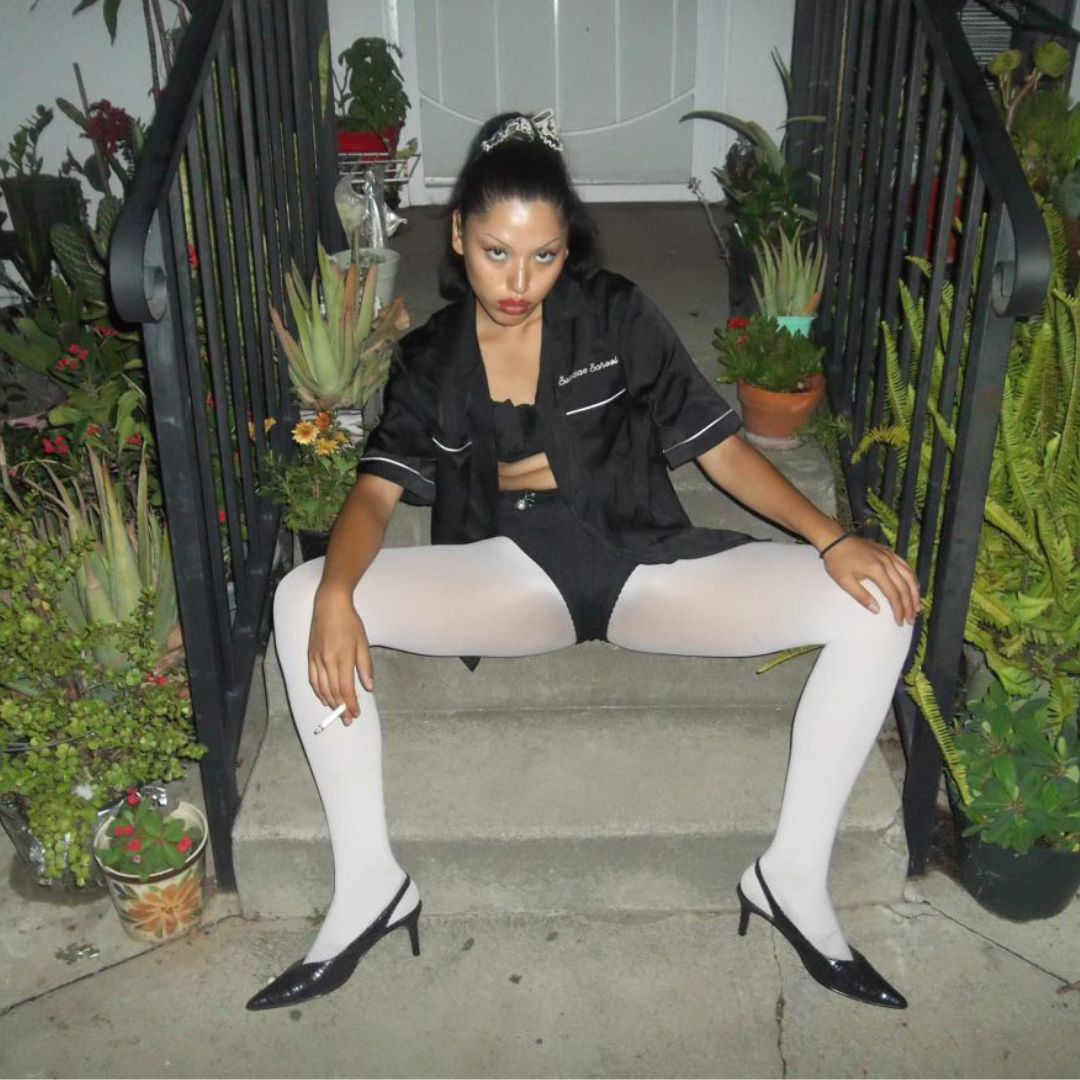 Seated on outdoor steps surrounded by plants, this person exudes edgy attitude. They wear a black bowling-style shirt over a bra top and short shorts, paired with white opaque tights and black pointed heels. A cigarette dangles from one hand and their bold makeup and dramatic pose add to the striking visual impact.