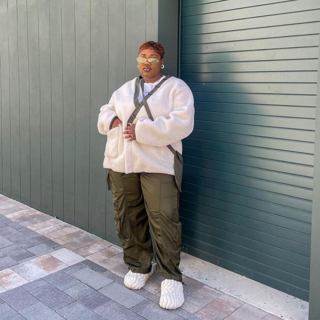 A stylish person stands confidently on a paved sidewalk in front of a forest green wall. They wear a cream sherpa fleece jacket, olive green ruched cargo pants, and textured off-white slip-on shoes. Accessories include angular sunglasses and a crossbody bag.