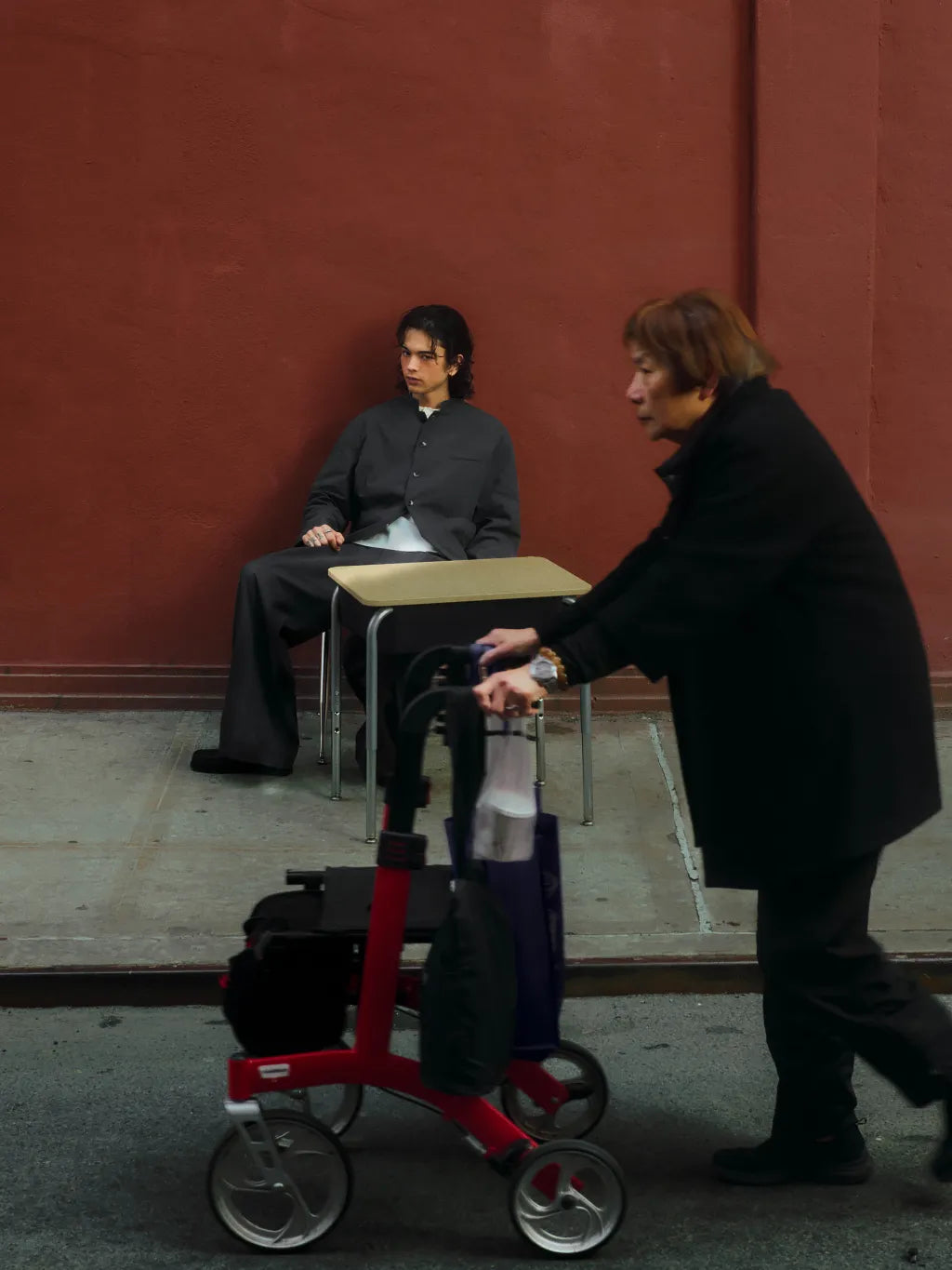 An old lady passing in front of a male model sitting in front of a school desk against a red wall wearing SUNDAE SCHOOL STAND COLLAR UNIFORM JACEKET and PLEATED UNIFORM PANTS