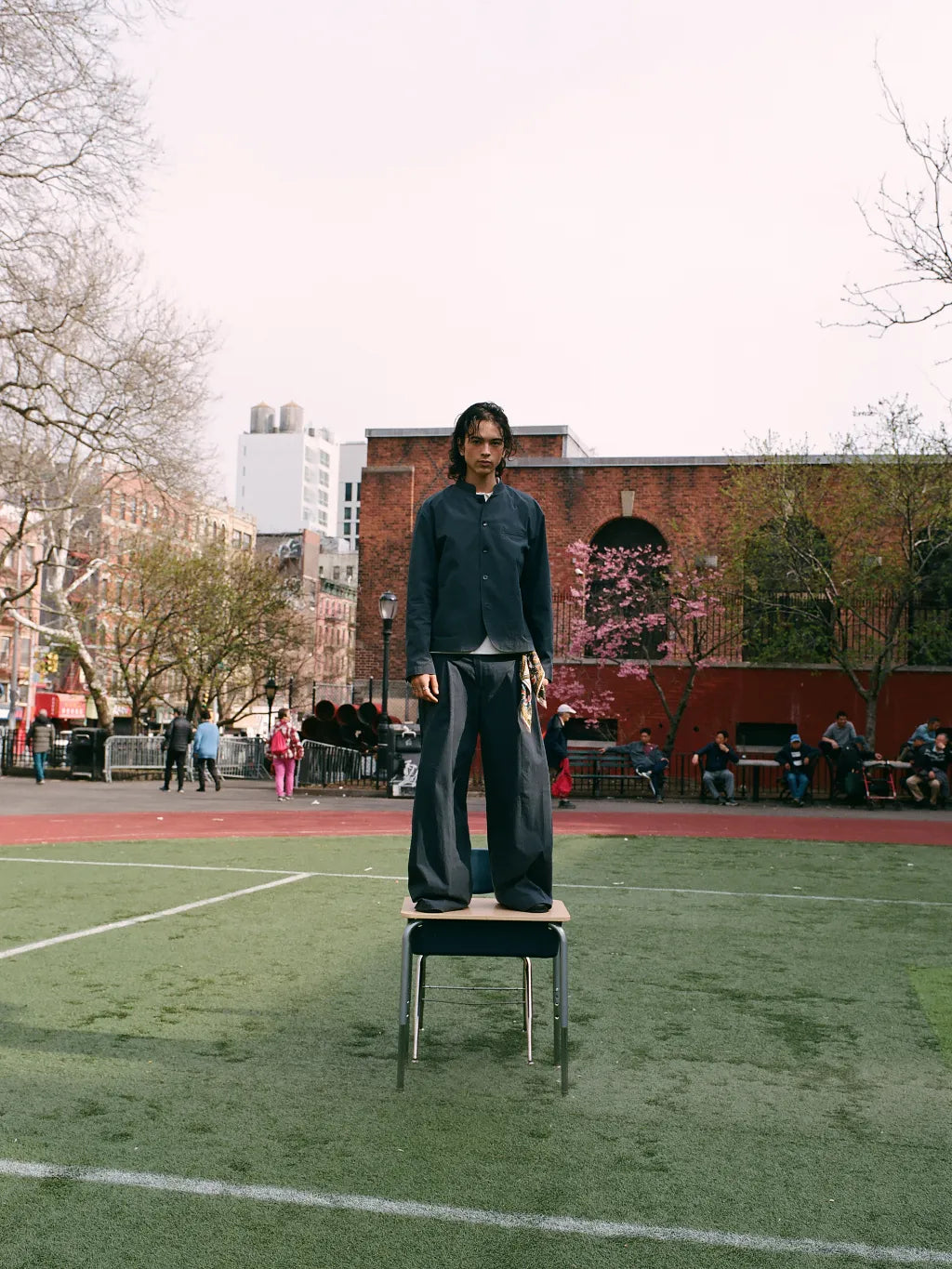 A person wearing SUNDAE SCHOOL STAND COLLAR UNIFORM JACKET and matching PLEATED UNIFORM PANTS stands on a small table in an outdoor urban park or sports field, with brick buildings, trees, and seated people in the background.