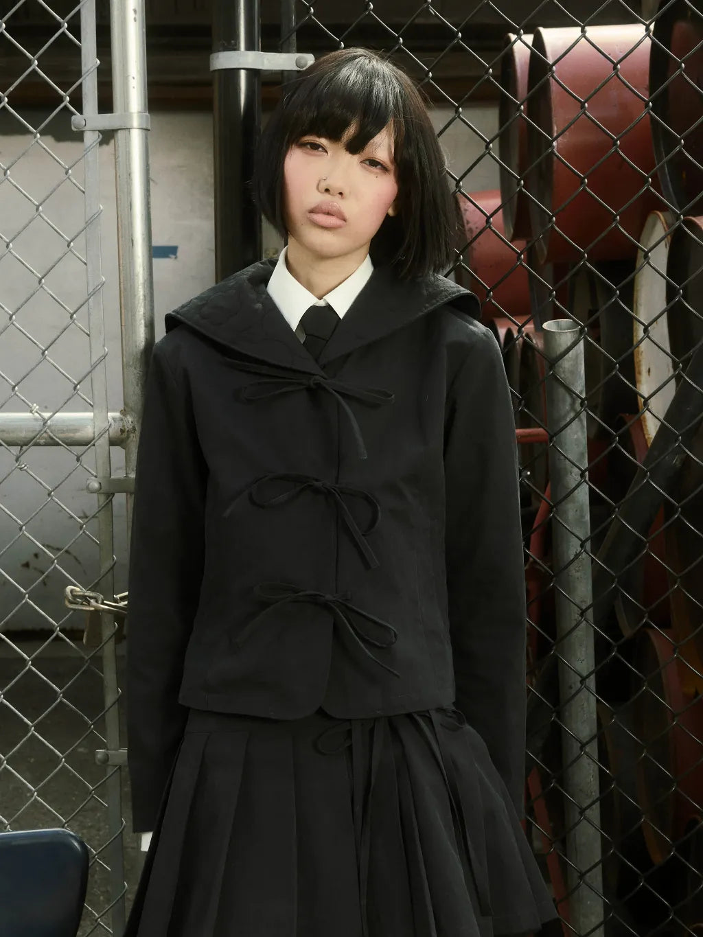A model wearing SUNDAE SCHOOL SAILOR COLLAR UNIFORM JACKET with tie details and a matching APRON SKIRT, layered over a white collared shirt and black tie, standing in front of a chain-link fence.