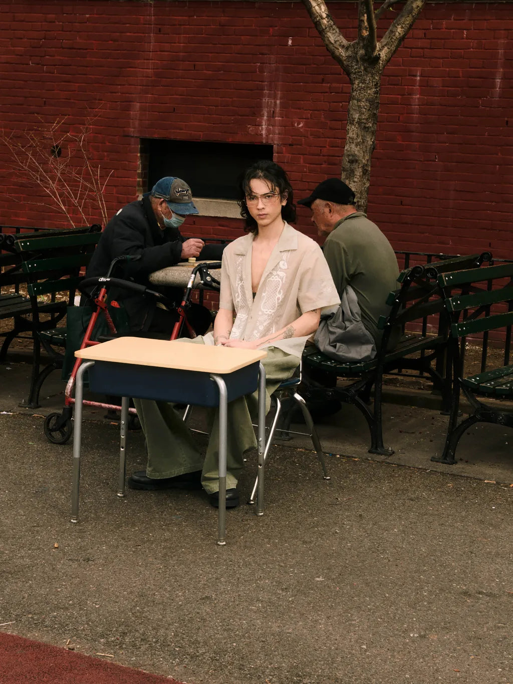 A person wearing SUNDAE SCHOOL BARONG SHIRT and OLIVE HANBOK CHINO PANTS sits at a small school desk in an outdoor urban setting with a red brick wall, park benches, and two men in the background.