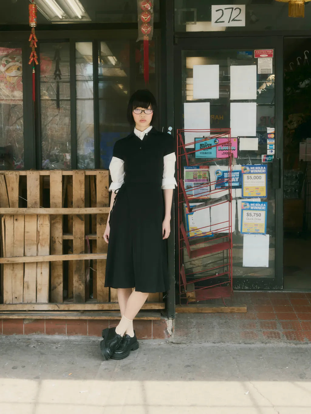 A woman wearing CINDY POLO DRESS over a white long-sleeve collared shirt stands in front of a storefront with a glass door, newspaper rack, and wooden pallet barriers.