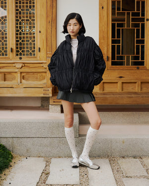 A woman with short dark hair and a neutral expression stands outdoors by a wooden building, wearing the Sundae School BLACK PLEATED DUVET PUFFER, a gray pleated mini-skirt, white knee-high socks with black dots, and silver shoes.