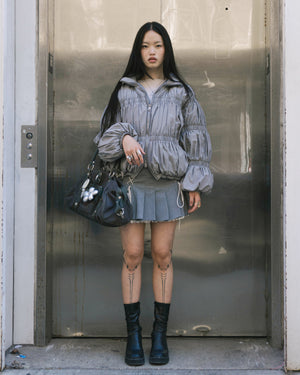 A woman with long straight hair and visible leg tattoos stands by an elevator in the SMOKE PLEATED DUVET PUFFER by Sundae School, a gray pleated mini skirt, black boots, and holding a large black handbag.
