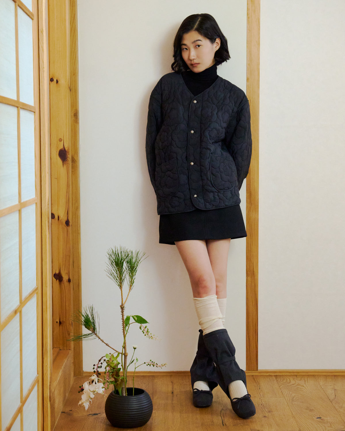 A woman with short black hair stands in a wooden room by a shoji screen, wearing the Sundae School BLACK MOCHI QUILTED MILITARY LINER, a black skirt, and white socks with dark sandals. A black vase with a pine branch and flowers sits beside her.