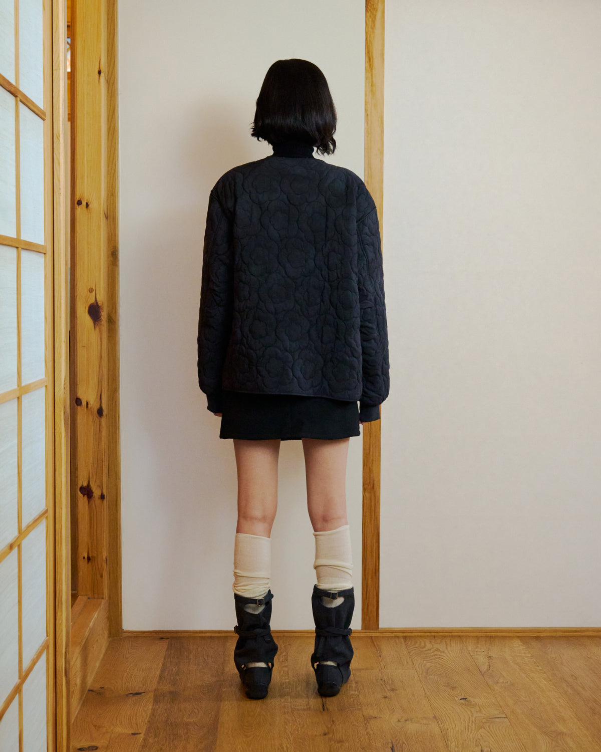 A person with short dark hair, back to the camera, stands in a wooden room facing a wall, wearing the Sundae School BLACK MOCHI QUILTED MILITARY LINER, a black skirt, cream socks, and black heeled shoes.
