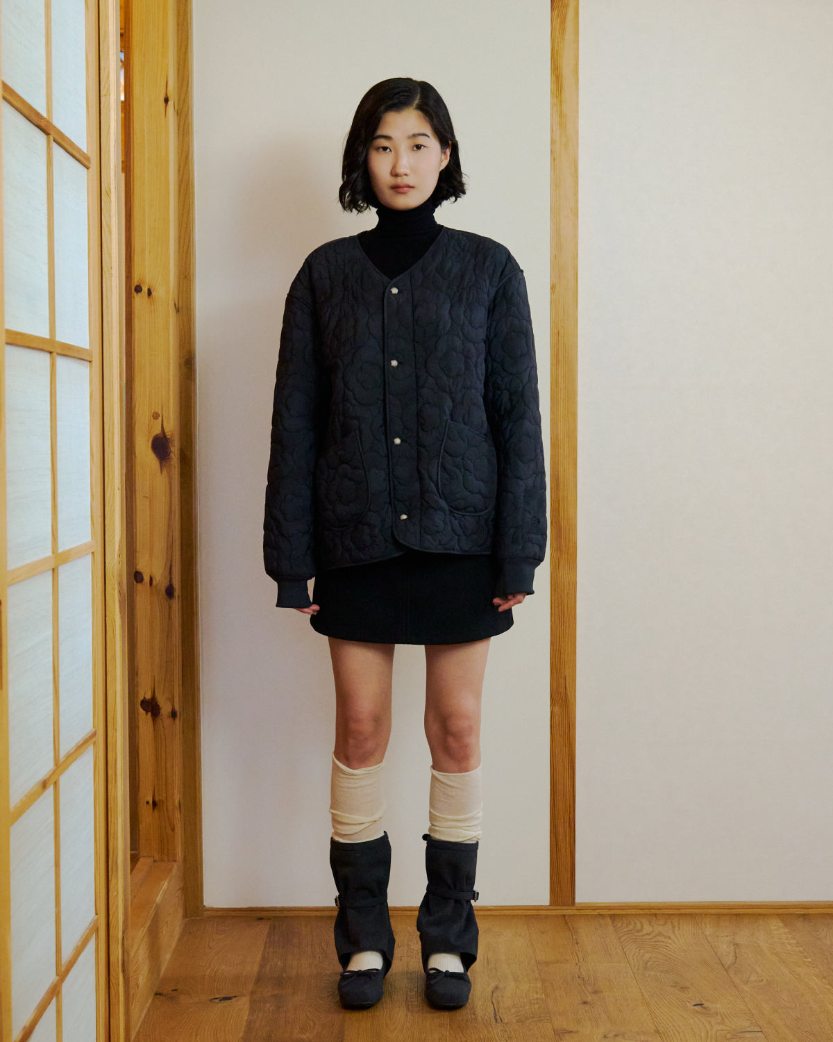 A young woman stands indoors on a wooden floor beside sliding wooden doors and light walls, wearing the Sundae School BLACK MOCHI QUILTED MILITARY LINER, a black turtleneck and skirt, tall black boots, and cream leg warmers.
