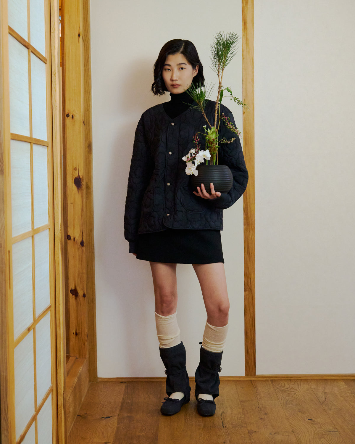 A woman stands indoors on a wooden floor, wearing the BLACK MOCHI QUILTED MILITARY LINER by Sundae School, paired with a black skirt, black boots, and cream leg warmers. She holds a potted plant against a plain, light-colored wall.