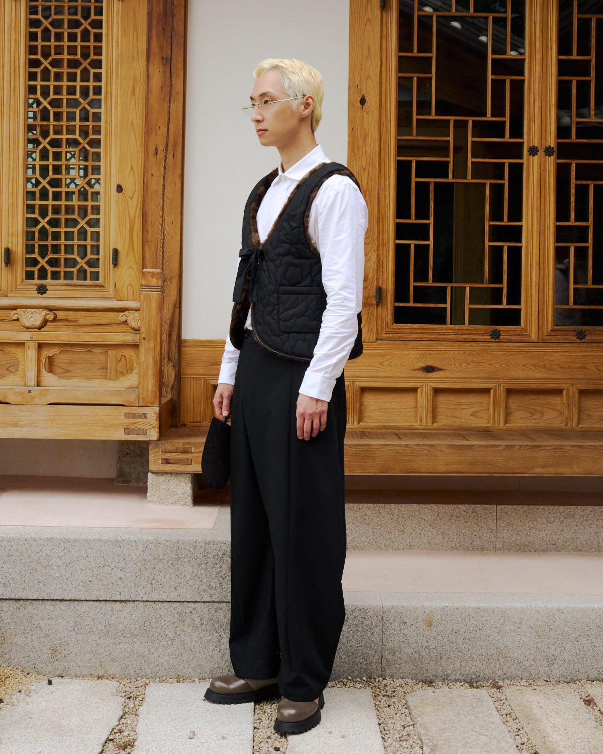 A person with short blond hair, glasses, and light skin stands on stone steps in front of a wooden building, wearing a white shirt, loose black pants, brown shoes, and Sundae School’s BLACK LEOPARD BAEJA VEST / BAG.