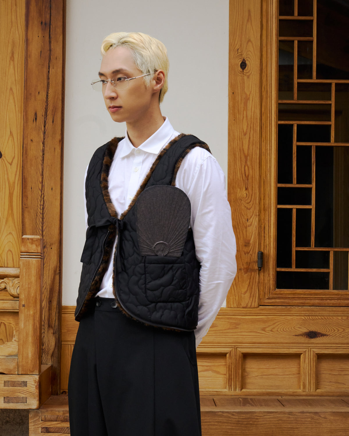 A person with short blond hair and glasses wears a white shirt, black pants, and the BLACK LEOPARD BAEJA VEST / BAG by Sundae School while standing in front of a carved wooden door with a geometric window.