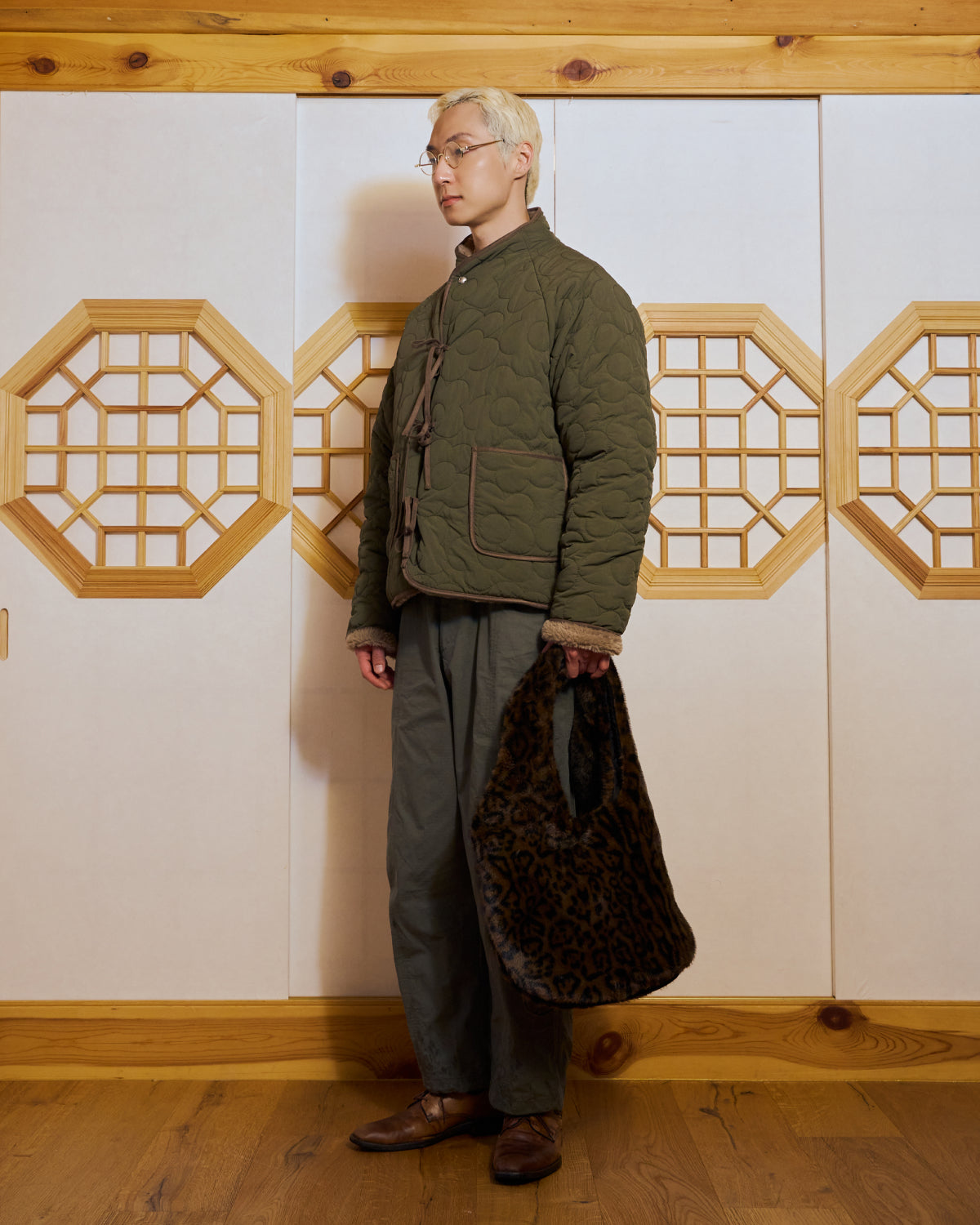 A person with short blond hair and glasses stands indoors against wooden paneling and geometric windows, wearing Sundae School’s olive green Korean quilted jacket, gray pants, brown shoes, and holding the BLACK LEOPARD BAEJA VEST / BAG.