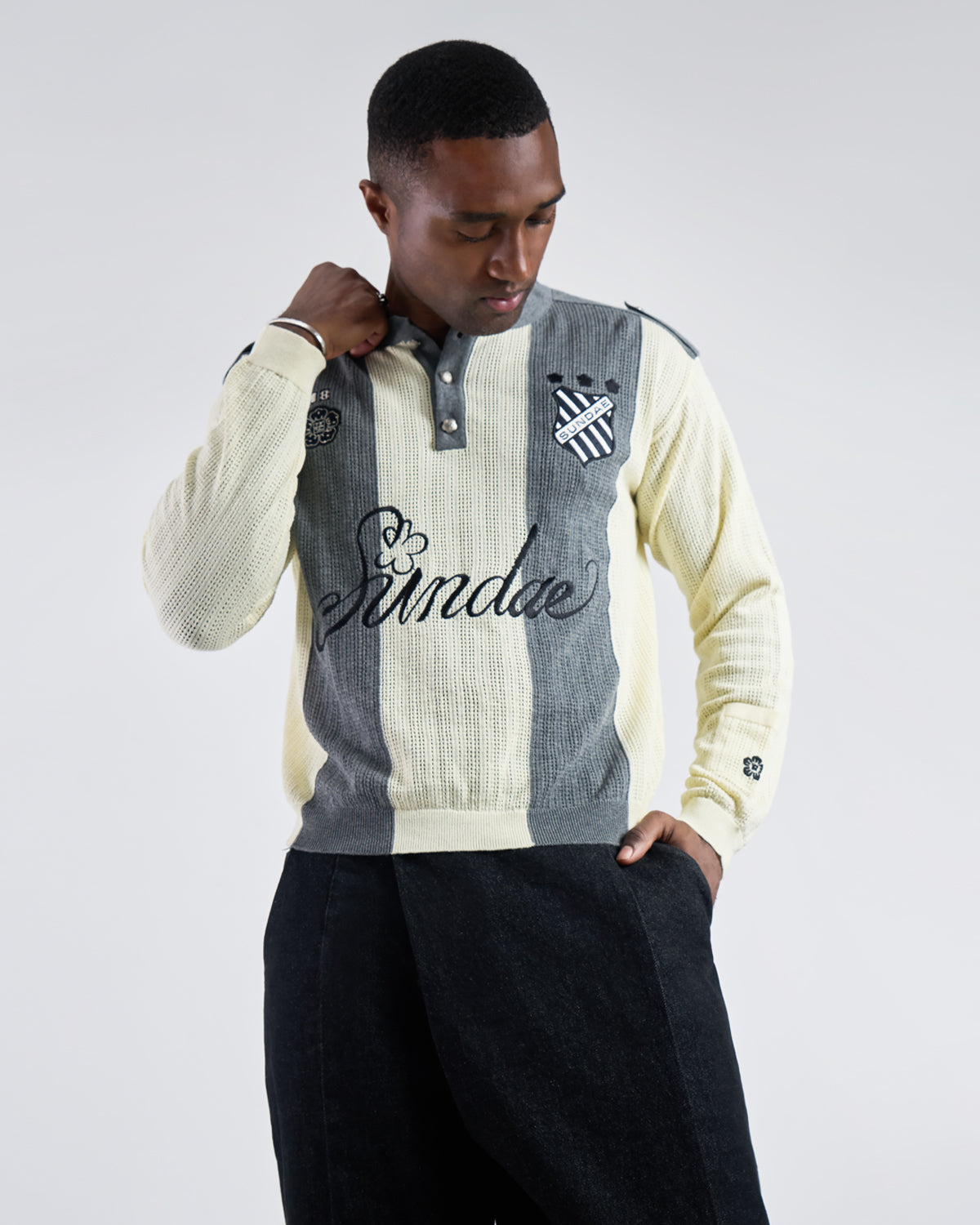 Butter Yellow Charcoal Sundae Soccer Jersey Knit modeled in casual lifestyle pose with model's hand raised to collar, showing vertical stripe pattern, henley placket, and embroidered details, styled with black pants, photographed against light background with natural lighting.