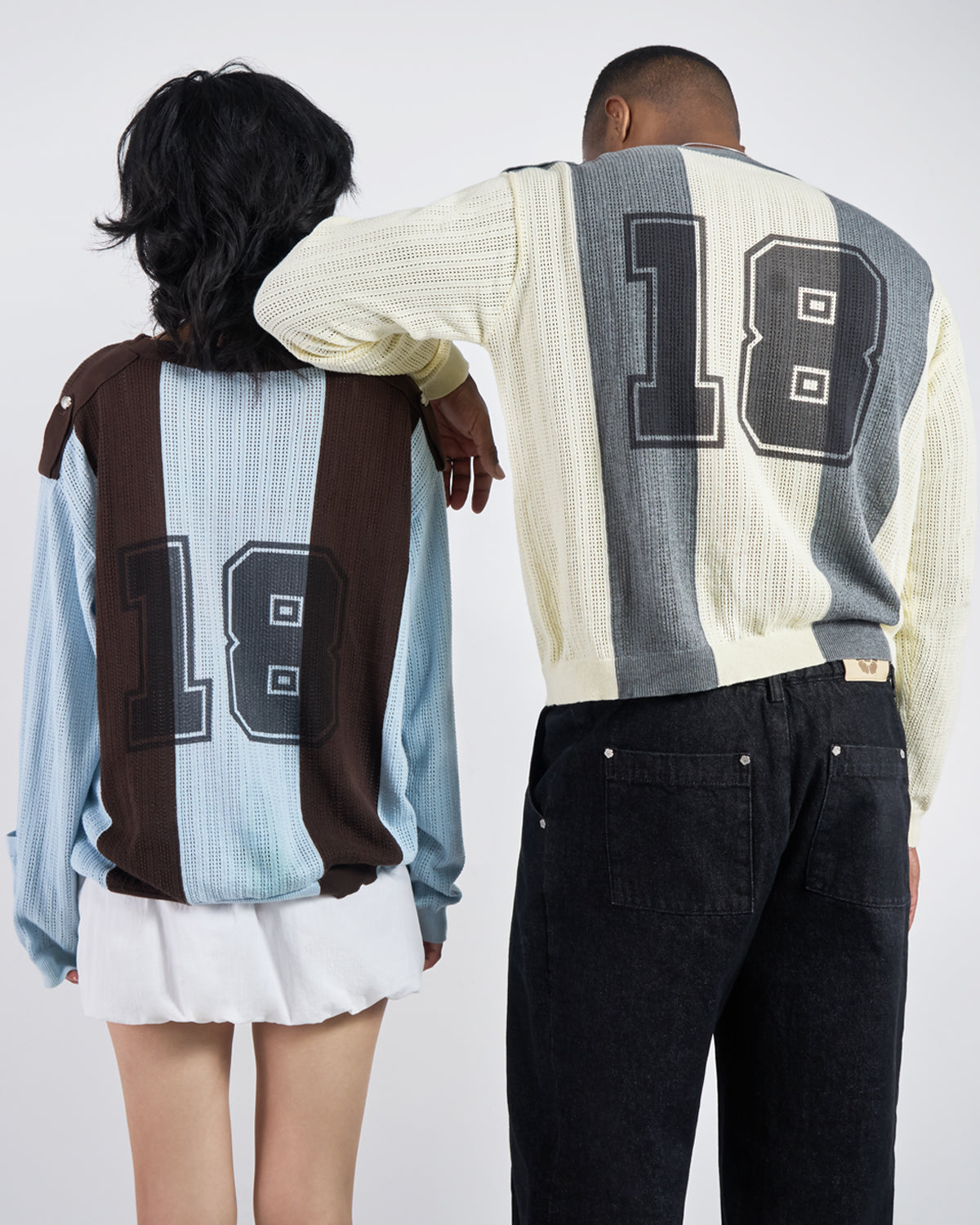 Two people stand side by side facing away, both in Sundae School BUTTER YELLOW CHARCOAL SUNDAE SOCCER JERSEY KNIT shirts with large 18s on the back—one with blue and brown stripes, the other in cream and gray.