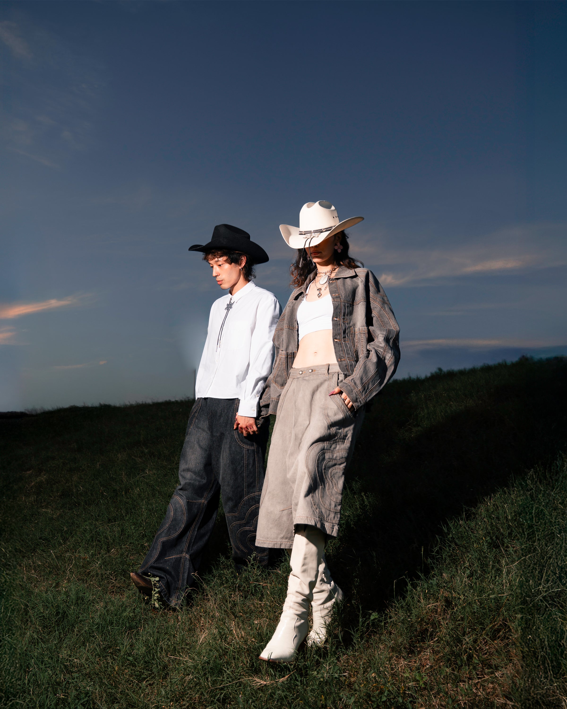 Two people in cowboy hats and modern western outfits walk hand in hand at sunset. One wears a white shirt paired with the BLACK MOCHI PANELED PANTS by Sundae School (x JOONIVERSE); the other sports a white top, long skirt, and boots.