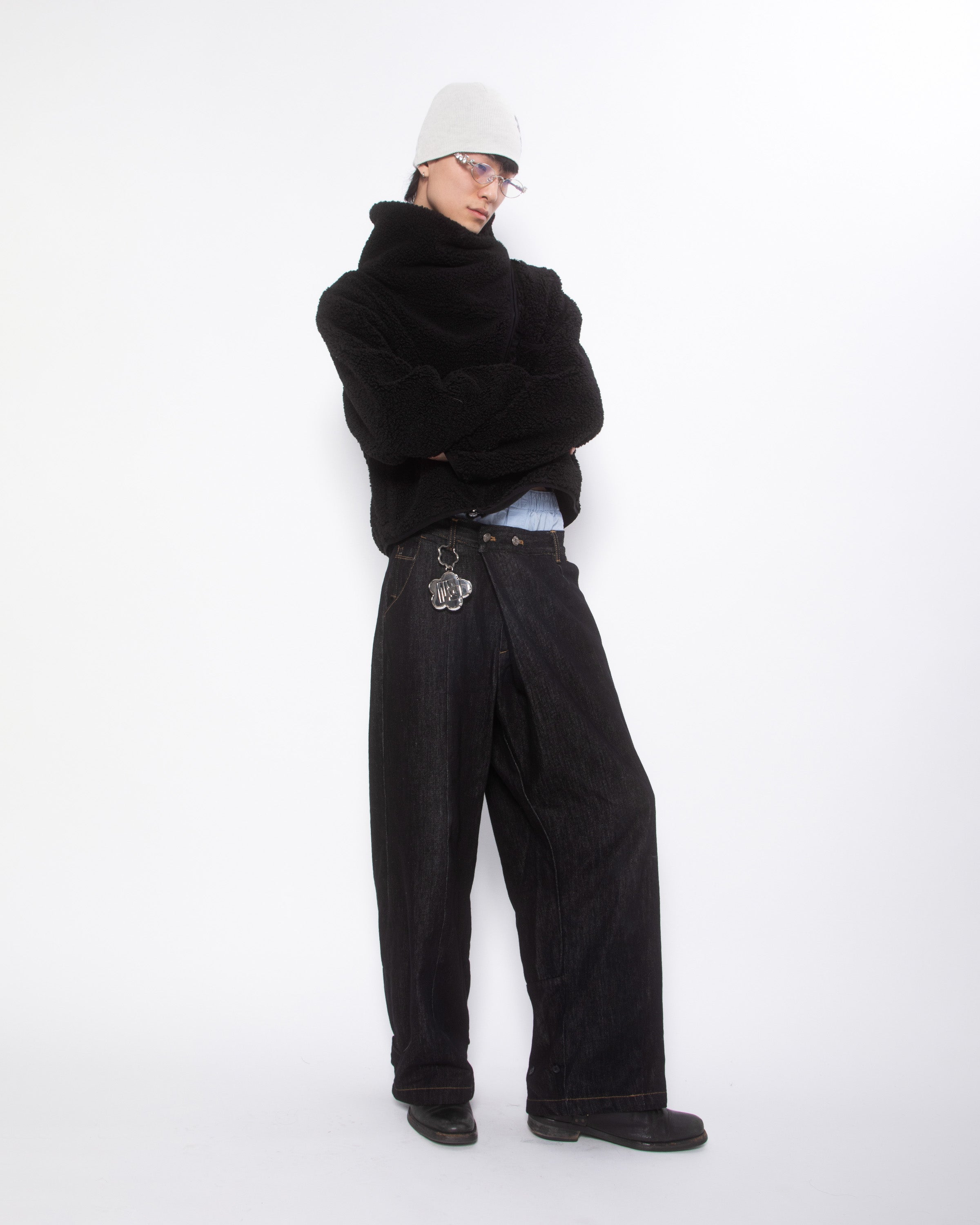 Close-up detail of Silver Mochi Lighter Case attached to black wide-leg jeans via flower-shaped keychain, person wearing black sherpa sweater and cream beanie, mochi case with Korean logo visible, white studio background.
